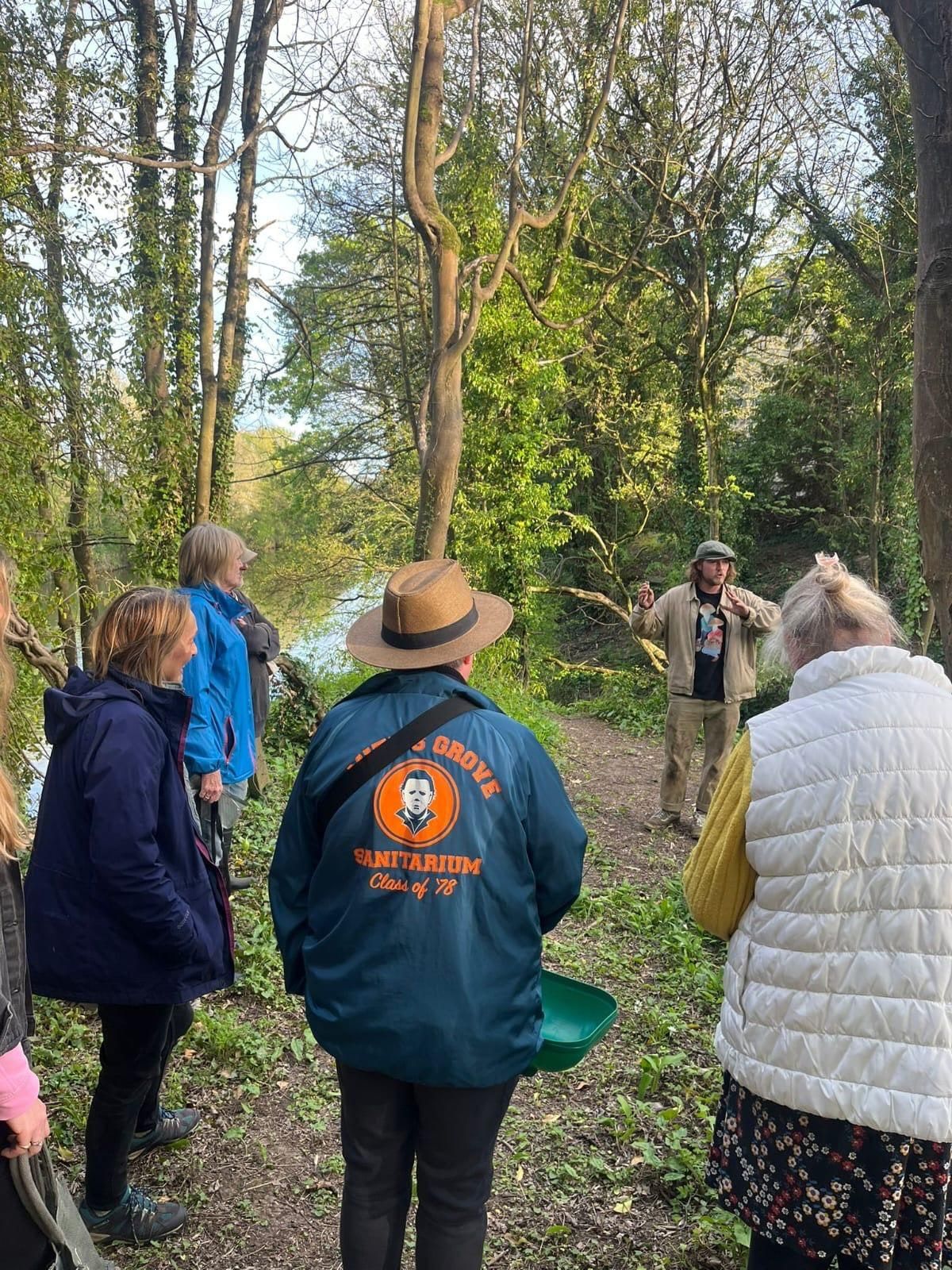 Willem, in a flat cap, talking to a small group on a wooded riverside path and gesturing at the trees around them.